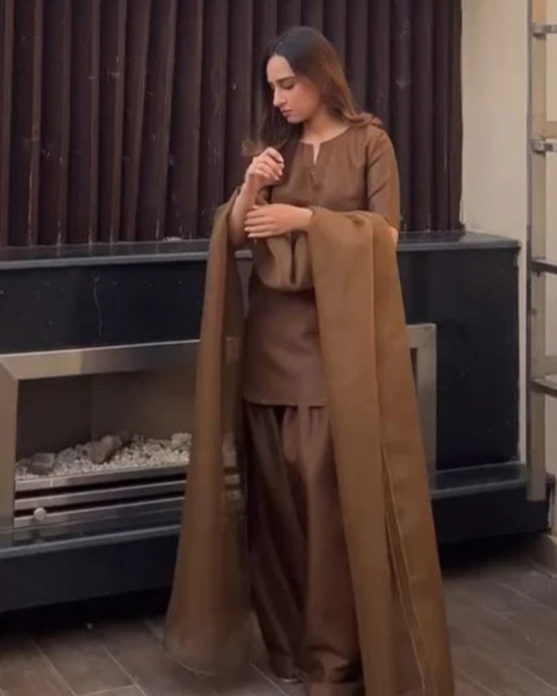 Woman in brown outfit standing by a fireplace outdoors. marhaba pret